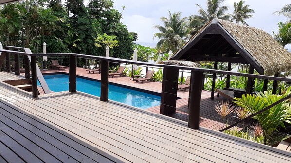 Um deck de madeira com piscina, espreguiçadeiras e uma estrutura com telhado de palha.