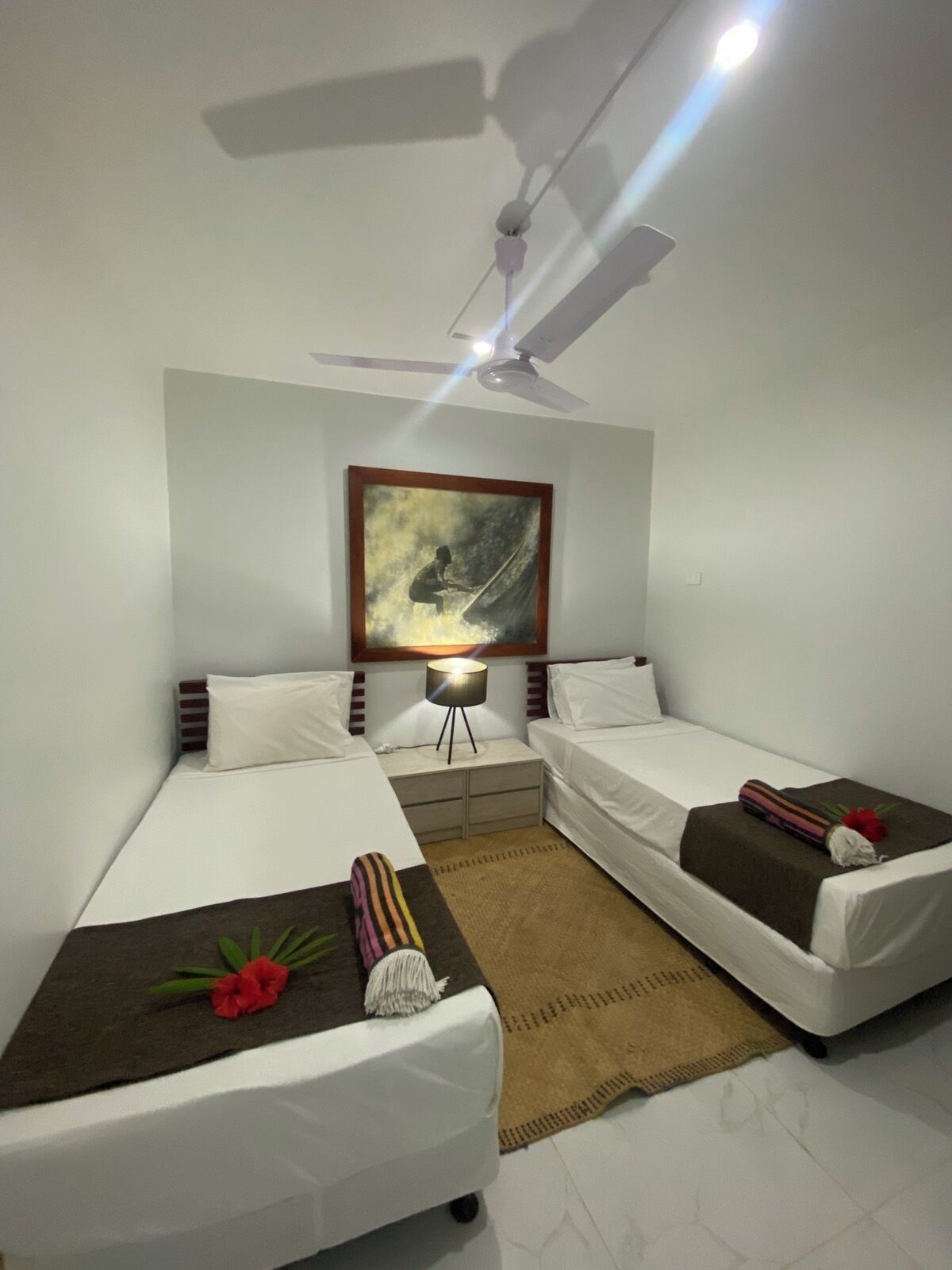 A hotel room with two single beds, a ceiling fan, a nightstand with a lamp, and a painting on the wall.