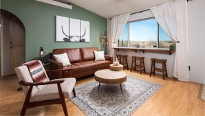 A living room with a brown leather sofa, a round wooden coffee table, and a patterned rug. There is a window with white curtains and a view of the desert.