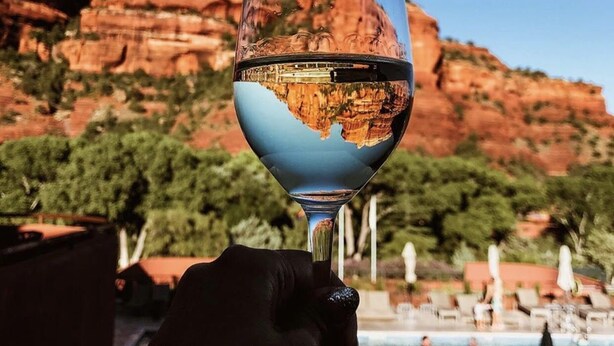 A glass of white wine held by a person with a reflection of a rocky landscape and a pool in the background.