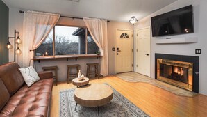 A cozy living room with a leather sofa, a fireplace, a TV mounted above it, and a window with curtains.