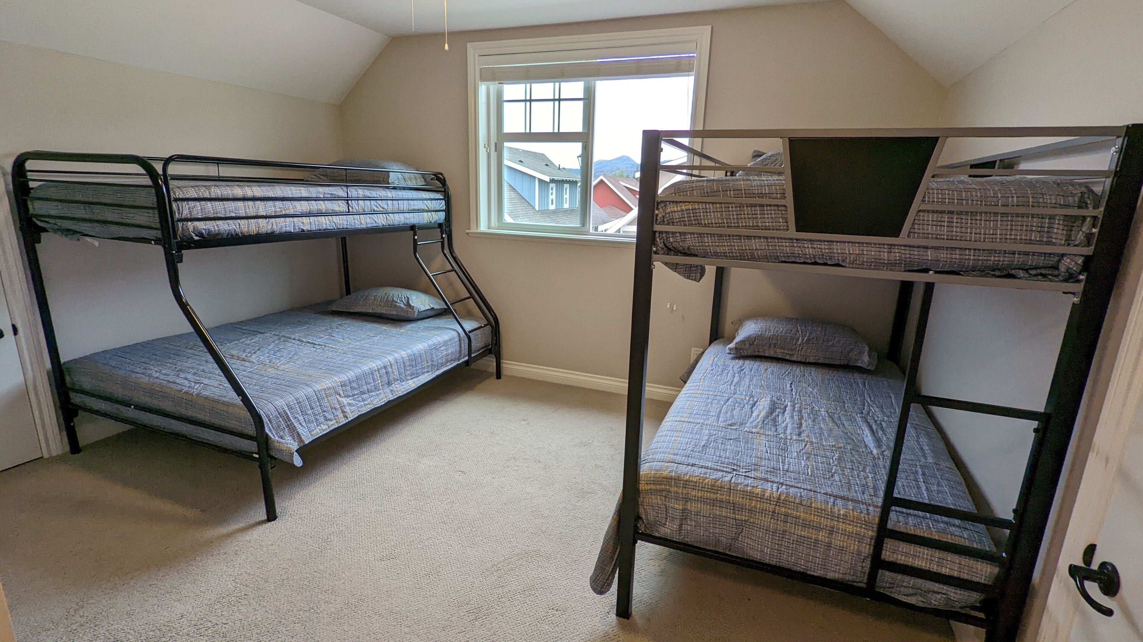 A room with two bunk beds, a window with a view of houses, and a carpeted floor.