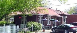 Ein Gebäude mit rotem Dach, weißen Fenstern und weißem Zaun. Draußen stehen Tische und Stühle, und ein Auto ist vor der Tür geparkt.