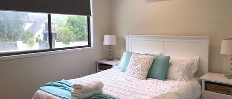 Un dormitorio con cama, mesita de noche, lámpara y ventana con vista a la vegetación exterior.