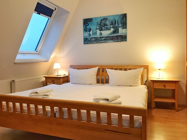 A bedroom with a sloped ceiling, a bed with white linens, two bedside tables with lamps, a radiator, and a painting on the wall.
