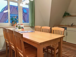 A dining area with a wooden table and chairs, a window with a view of a house, and a kitchen area with a sink and cabinets.