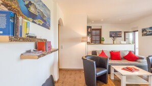 Ein modernes Wohnzimmer mit einer Couch, Sesseln und einem Couchtisch. Ein Bücherregal mit Büchern und ein Gemälde von einer Küstenlandschaft sind ebenfalls vorhanden.