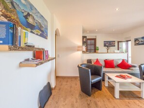 Ein modernes Wohnzimmer mit einer Couch, Sesseln und einem Couchtisch. Ein Bücherregal mit Büchern und ein Gemälde von einer Küstenlandschaft sind ebenfalls vorhanden.