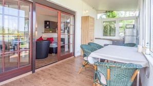Une cuisine moderne dotée d’un four et d’un micro-ondes encastrés, un coin repas avec des chaises en osier, et une porte-fenêtre coulissante donnant sur un espace extérieur aménagé pour se détendre.