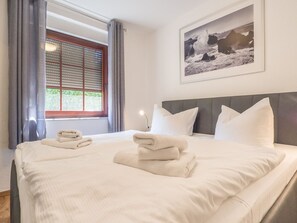 Ein ordentlich bezogenes Bett mit weißen Leinentüchern und Handtüchern, ein großes Fenster mit Jalousien und ein gerahmtes Foto an der Wand.