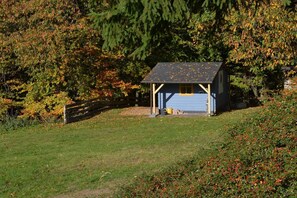 Une petite cabane à un seul étage, dotée d’un toit à pignon, entourée d’une clôture en bois et d’un feuillage dense.
