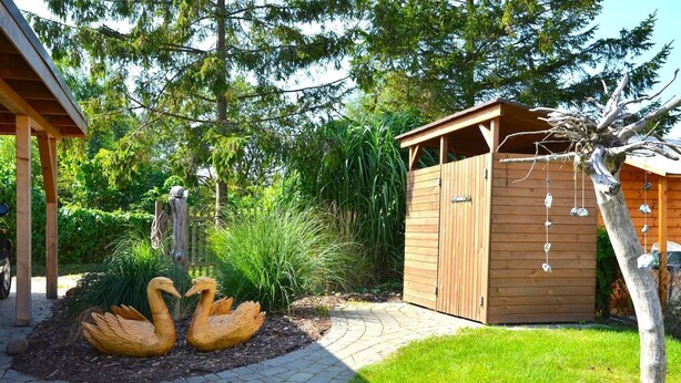 A wooden shed with a slanted roof, a paved pathway leading to it, and two swan sculptures in the foreground. - Vacation home "Lütt Eiland" (Glowe)