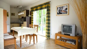 A dining area with a table set for two, a kitchen in the background, a television on a wooden stand, and a window with curtains. - Vacation home "Lütt Eiland" (Glowe)