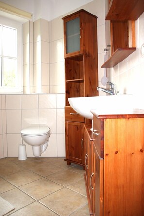 A bathroom with a toilet, sink, and wooden cabinets. - Vacation home "Lütt Eiland" (Glowe)