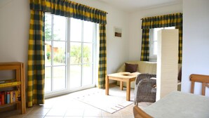 A room with a large sliding glass door, a wooden table, a wicker chair, and a bookshelf. - Vacation home "Lütt Eiland" (Glowe)