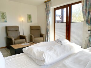 Une chambre d’hôtel avec deux fauteuils en cuir beige, une table basse en bois avec une télécommande, un lit blanc avec une couverture soigneusement pliée et une fenêtre avec des stores.