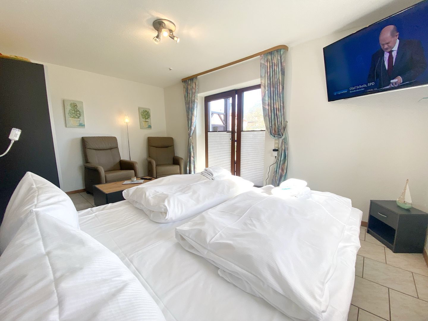 Ein Hotelzimmer mit Bett, zwei Stühlen, einem Fernseher und einem Fenster mit Vorhängen.