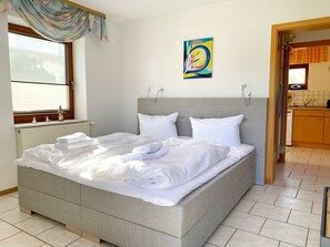 A neatly made bed with white linens and towels, a grey headboard, and a window with blinds.