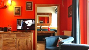 A living room with a TV, wooden cabinets, a blue armchair, and a kitchen area visible through an open doorway.