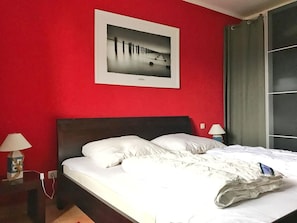 A hotel room with a red wall, a framed photograph, a bed with white bedding, a nightstand with a lamp, and a window with curtains.