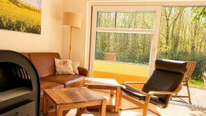 A living room with a sofa, a wooden coffee table, a leather chair, a lamp, and a large window with a view of trees. - Vacation home Emma - Apartment 2 (Lohme)