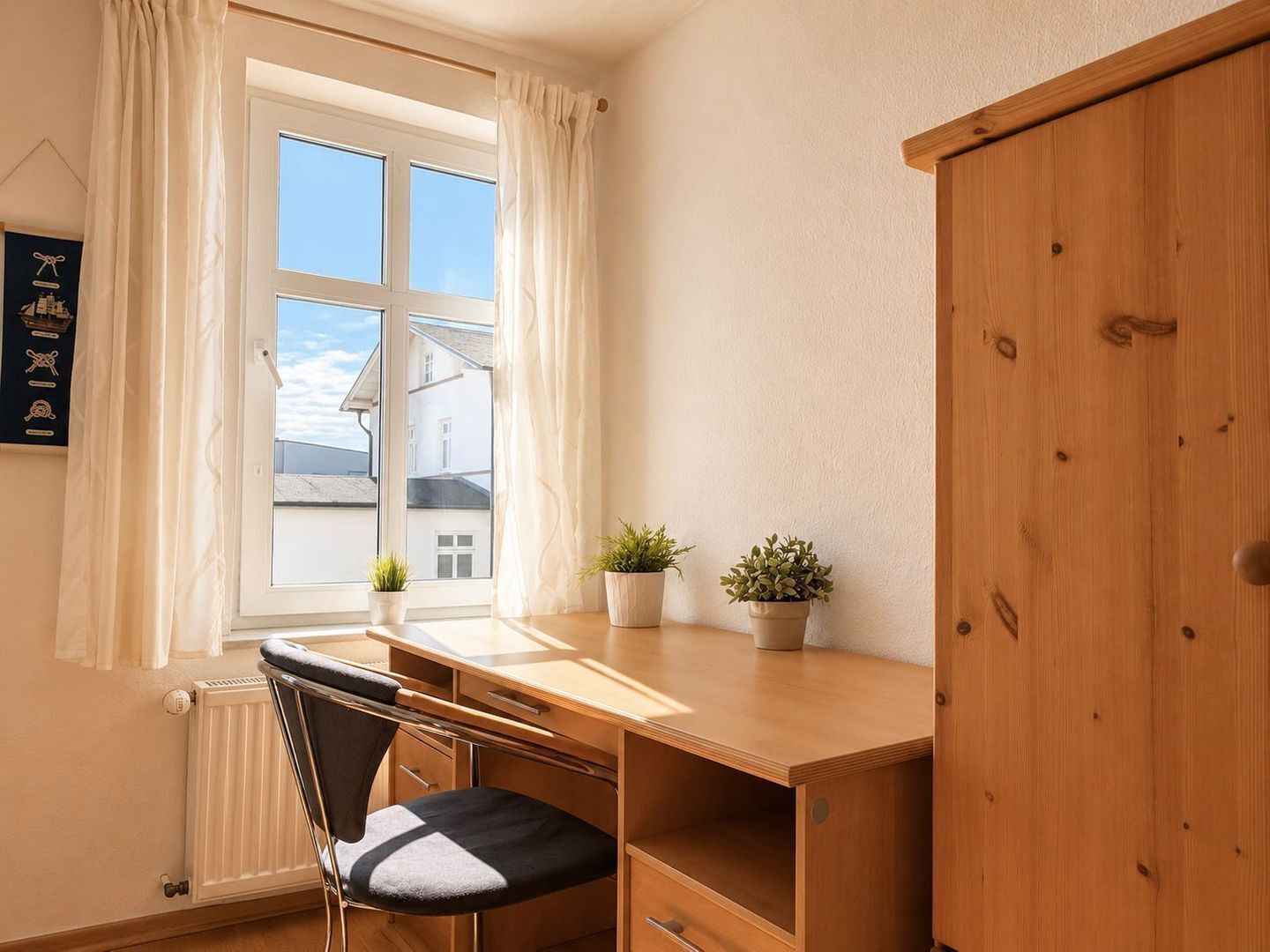 Ein Zimmer mit Schreibtisch, Stuhl, Heizkörper, einem Fenster mit Vorhängen und einem Schrank aus Holz.