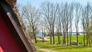 Uma varanda com mesa e cadeiras, um vaso de flores e vista para um parque com árvores e um corpo d'água.