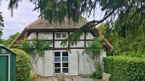 A traditional house with a thatched roof, white walls, and dark trim. In front, there is a small patio with a table and chairs. - Doppelhaushälfte Nr. 2 im Feriengarten (Nardevitz auf Rügen)