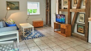 A cozy living room with a white sofa, a small wooden table, a TV on a wooden stand, and a window with a view of greenery. - Doppelhaushälfte Nr. 2 im Feriengarten (Nardevitz auf Rügen)