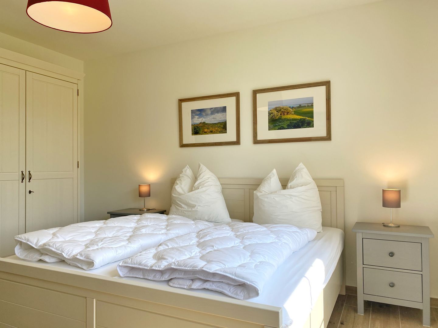 Un lit bien fait, avec une literie blanche, deux tables de chevet avec des lampes et deux tableaux encadrés accrochés au mur.