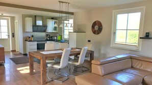 A modern kitchen with white cabinets, a dining table with chairs, a leather sofa, and a large window.