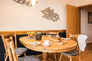 Une table de salle à manger ronde en bois, sur laquelle se trouvent un saladier de fruits, une plante en pot et du pain sur une planche à découper.