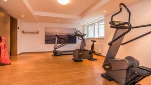 Um quarto amplo com piso de madeira, equipado com esteira elíptica, bicicleta ergométrica e esteira. Uma grande janela oferece luz natural, e uma pintura de uma cena de praia é visível na parede.