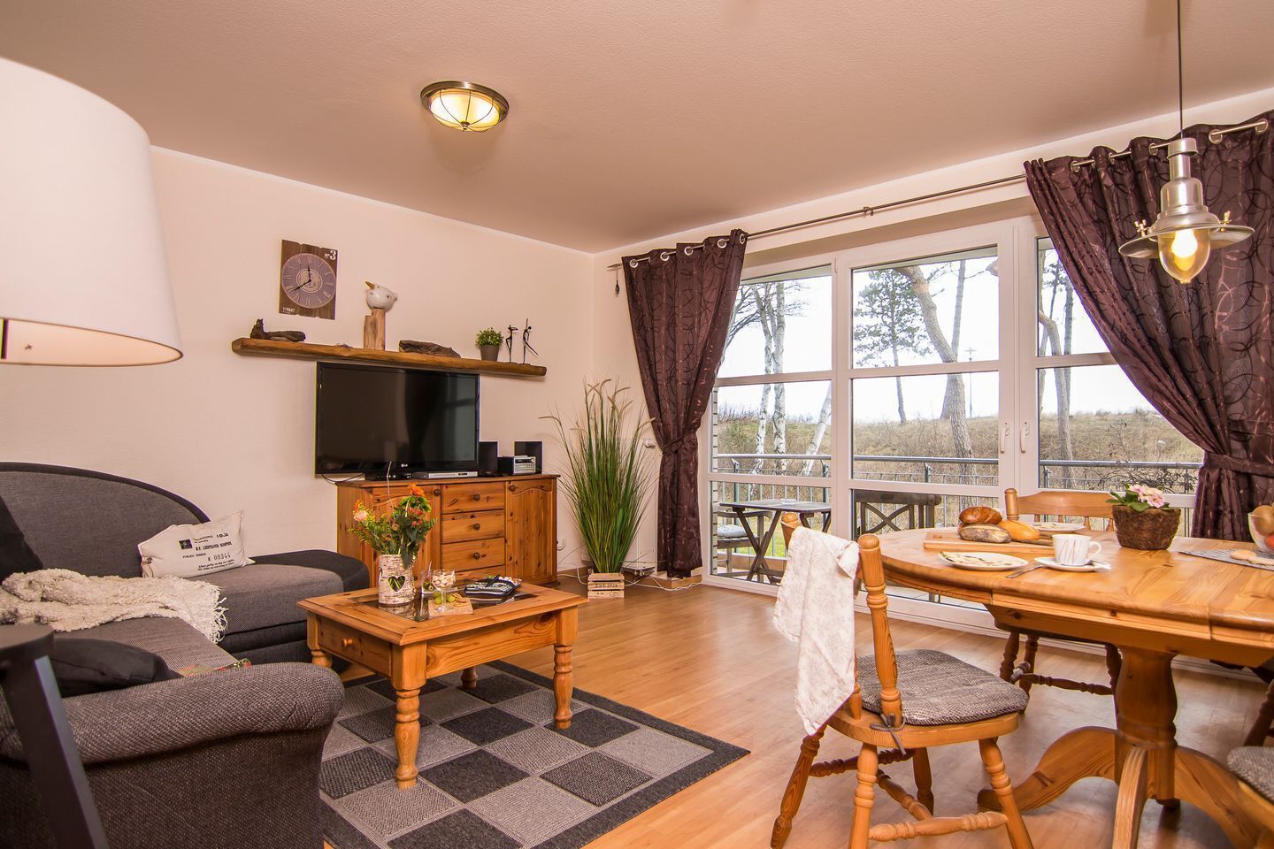 Een gezellige woonkamer met een bank, salontafel en televisie. Er is een eethoek met een tafel en stoelen, en een groot raam met gordijnen.