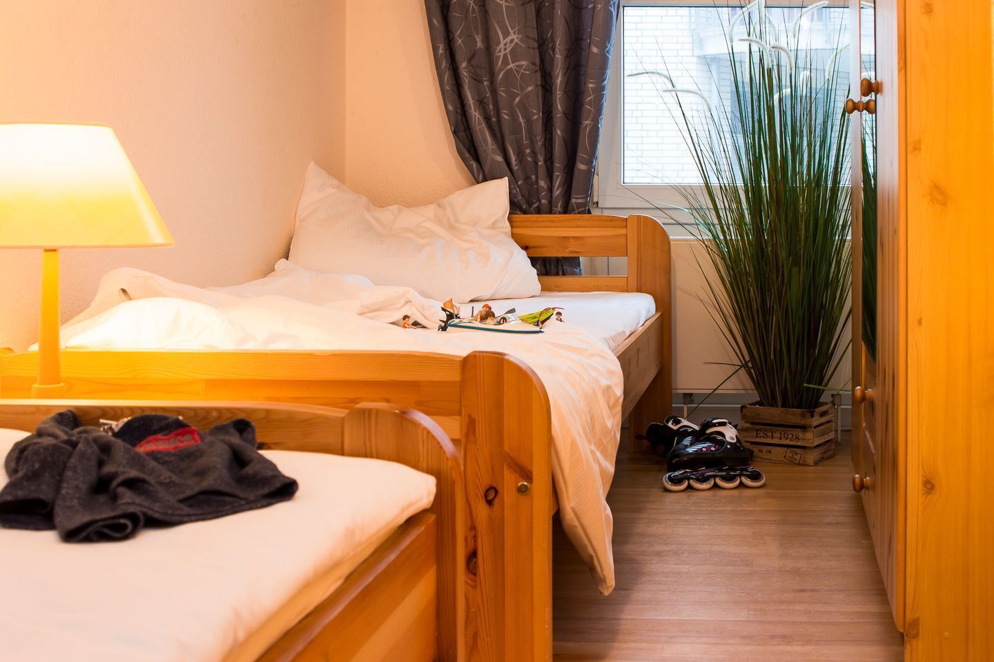 Een knus interieur met een houten bed, een nachtkastje met lamp, een klein plantje en een schoenenrek.