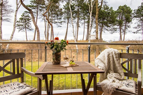 Une table en bois ornée d’un vase de fleurs, d’un verre de vin et d’une assiette de nourriture, sur un balcon avec des chaises en bois et une couverture.