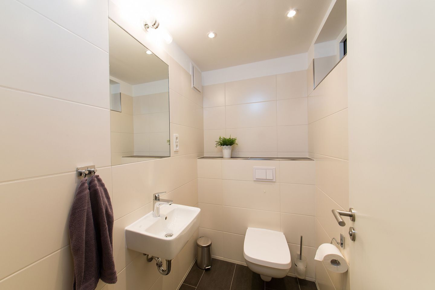 Ein modernes Badezimmer mit einem weißen Waschbecken, einer Toilette, einem Spiegel und einer kleinen Topfpflanze an der Wand.