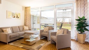 A modern living room with a sofa, armchairs, a glass coffee table, and a large window with curtains. - Dünenresort Binz - Apartment 4.1.6 (Binz)