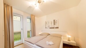 A bedroom with a large bed, two bedside tables with lamps, a window with curtains, and artwork on the wall. - Dünenresort Binz - Apartment 4.1.6 (Binz)