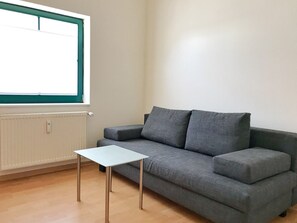 Una habitación con un sofá gris, una mesita blanca pequeña, una ventana con marco verde y un radiador.