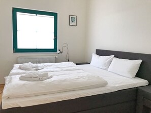 Una cama bien hecha con sábanas y almohadas blancas, una lámpara de noche, un cuadro enmarcado en la pared y una ventana con marco color aguamarina.