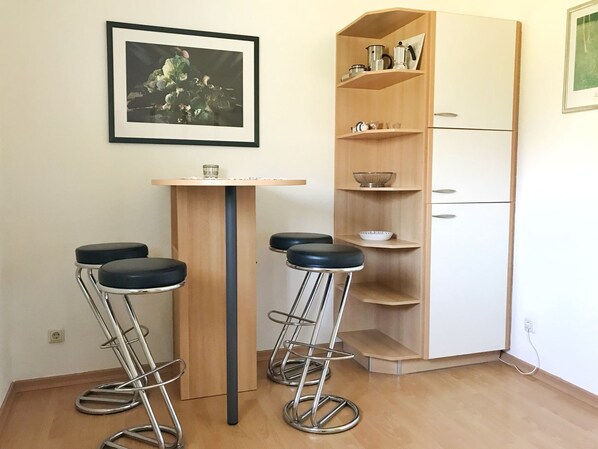 Una cocina con una mesa de madera, dos taburetes altos, una cafetera y un armario con estantes.