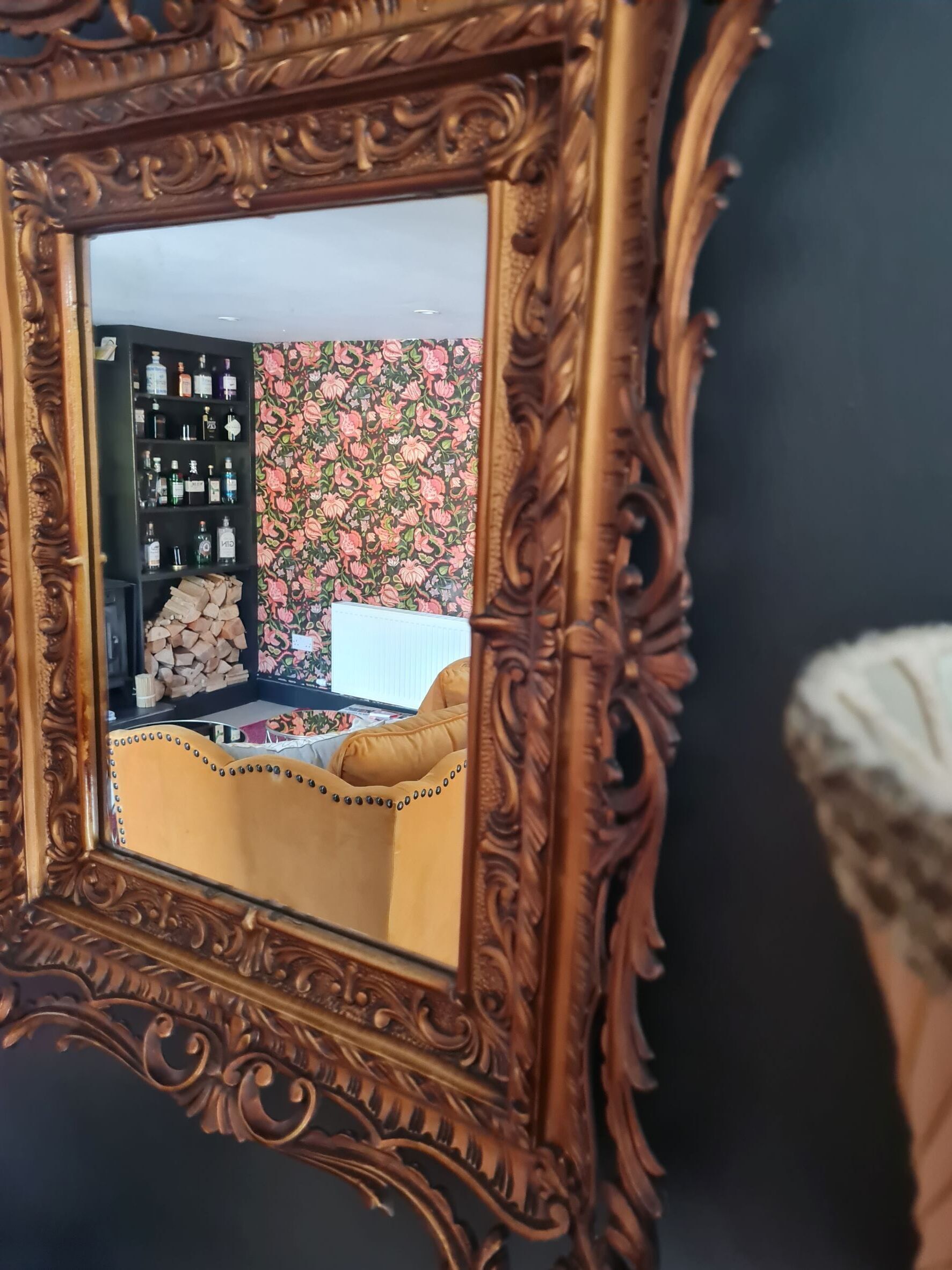 A mirror with an ornate frame reflecting a room with a floral wallpaper, a bookshelf, and a yellow sofa.