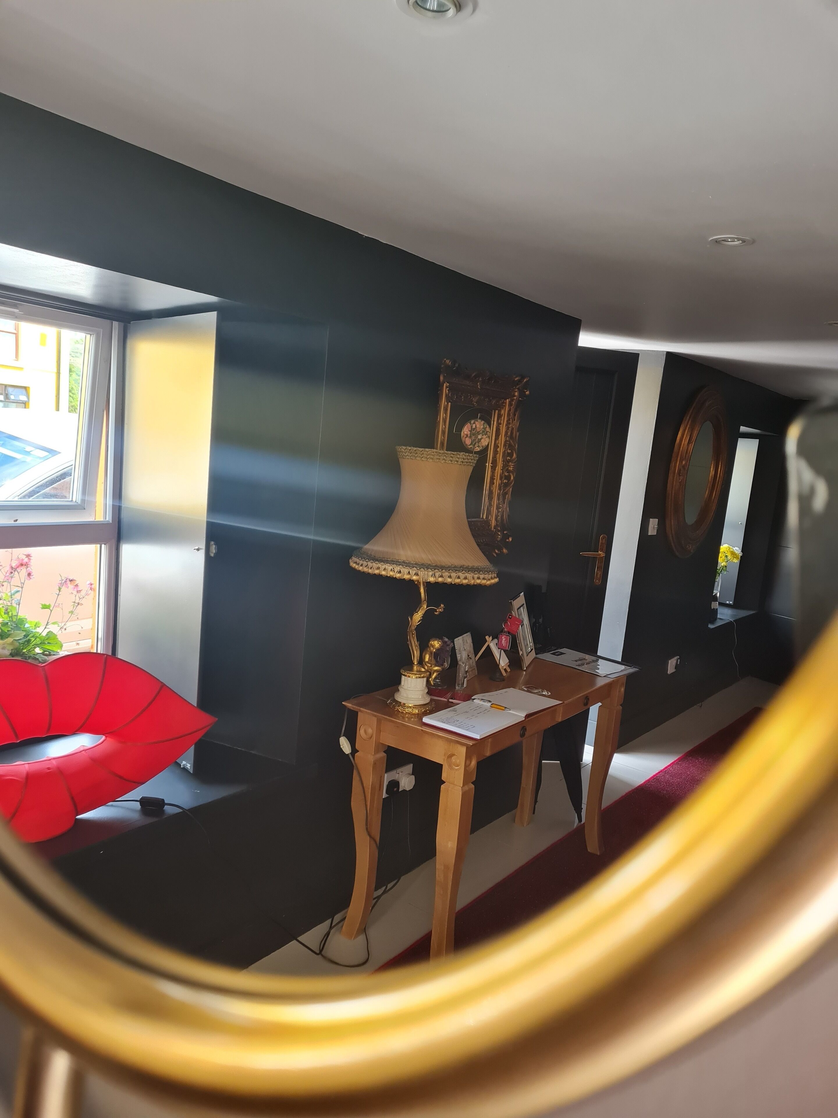 Una habitación con una pared oscura, una silla roja, una mesa de madera con una lámpara y un espejo.