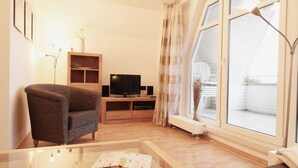 A living room with a brown armchair, a glass coffee table with a book, a TV on a wooden stand, and a large window with curtains.