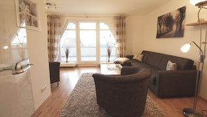 A modern living room with a sofa, armchairs, a coffee table, and a floor lamp. There is a large window with curtains, a painting on the wall, and a door with a handle.