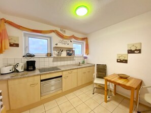 Una cocina con horno integrado, cafetera y lavamanos. Hay una ventana sobre el lavamanos y una estantería con utensilios de cocina.