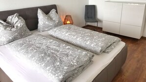 A modern bedroom with a grey upholstered bed, patterned bedding, a bedside lamp, a grey chair, and a white wardrobe. - Ferienappartement "langschläfer" (Sassnitz)