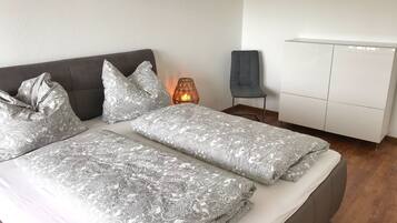 A modern bedroom with a grey upholstered bed, patterned bedding, a bedside lamp, a grey chair, and a white wardrobe.