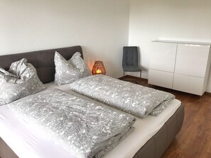Ein modernes Schlafzimmer mit einem grau gepolsterten Bett, geblümter Bettwäsche, einer Nachttischlampe, einem grauen Sessel und einem weißen Kleiderschrank.